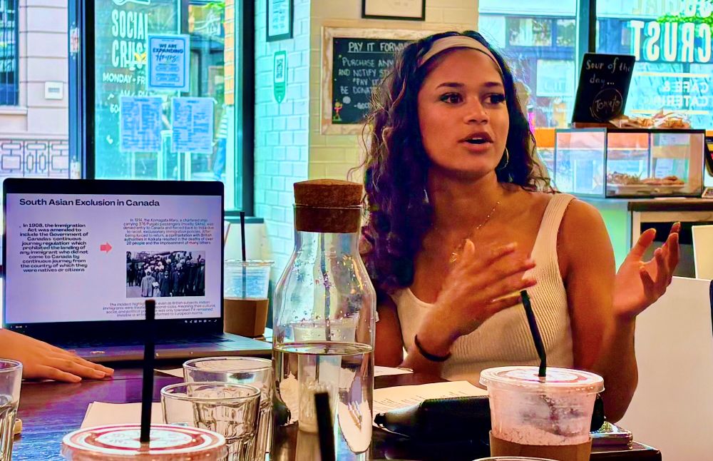 Zoya Reebye is seated at a crowded table in a coffee shop. She is gesturing with her hands, mid-speech. She has long dark wavy hair pulled back with a headband. She is wearing a light-coloured tank top.