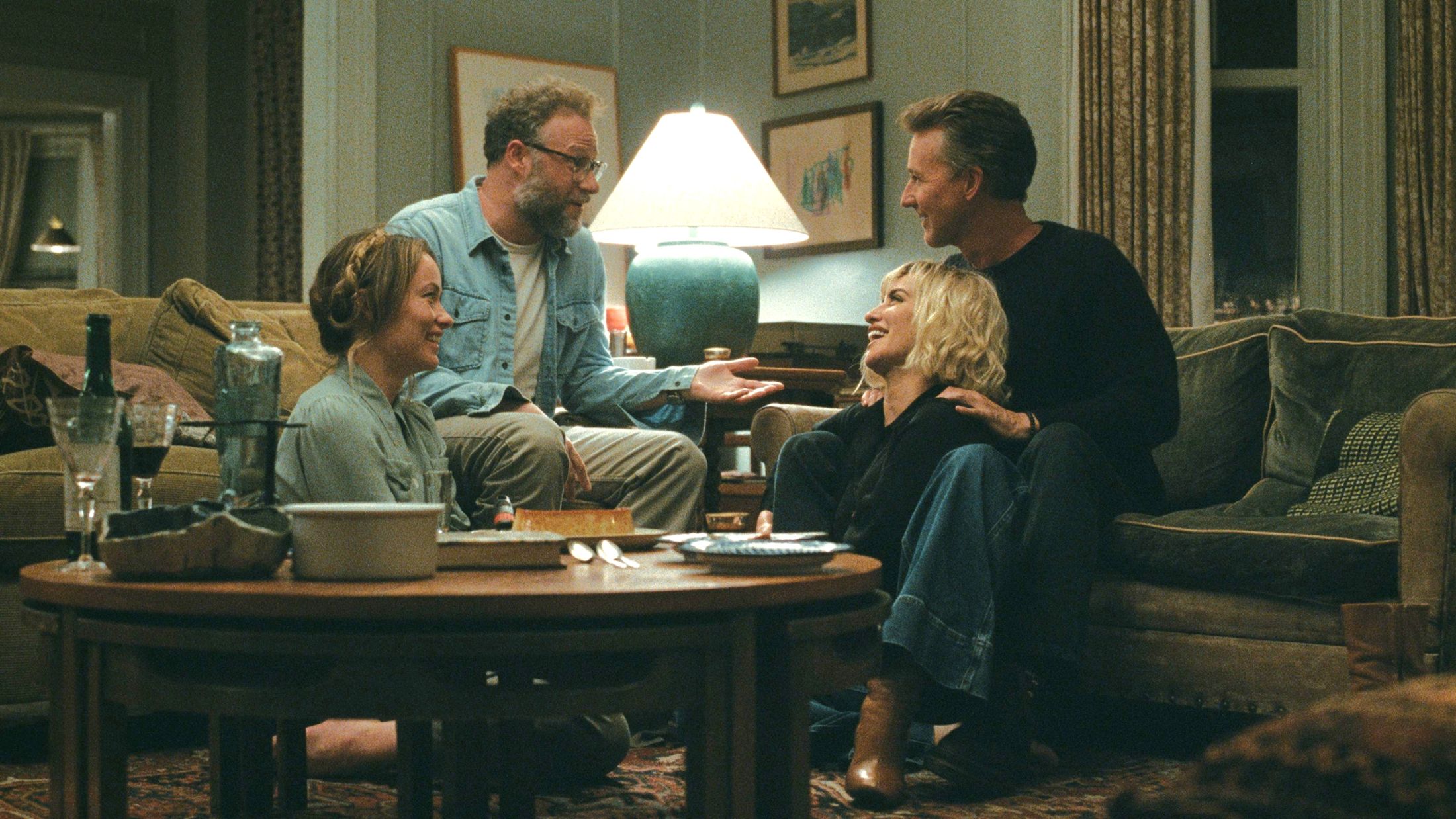 In a still of 'The Invite,' Seth Rogen, who plays Joe, and Olivia Wilde, who plays Angela, sit on the couch and floor across from Edward Norton, who plays Hawk, and Penelope Cruz, who plays Pina, laughing and talking. 
