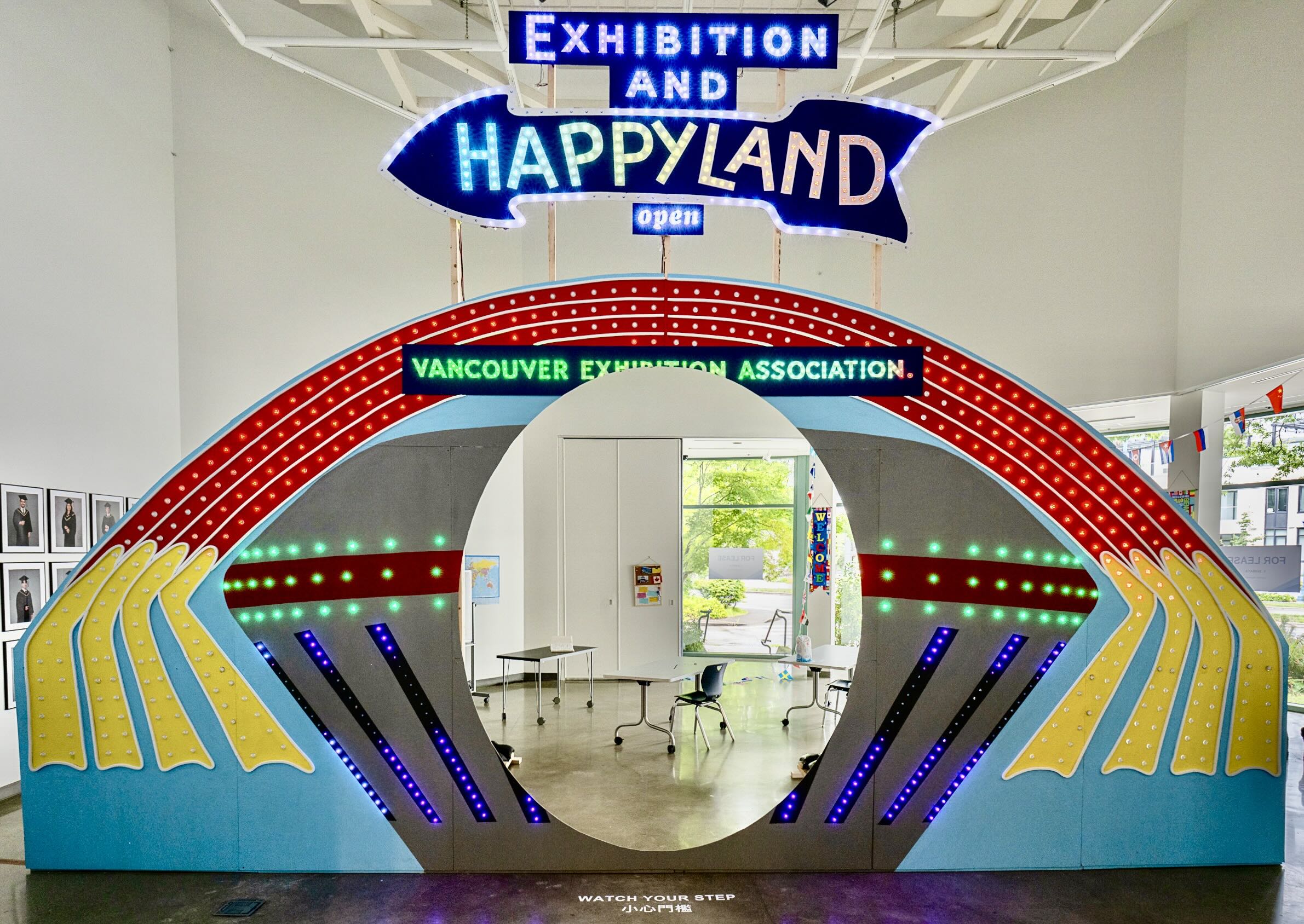 A large installation in a gallery space features a colourful, brightly lit tunnel above which a sign reads &ldquo;Exhibition and Happyland open; Vancouver exhibition association.&rdquo;