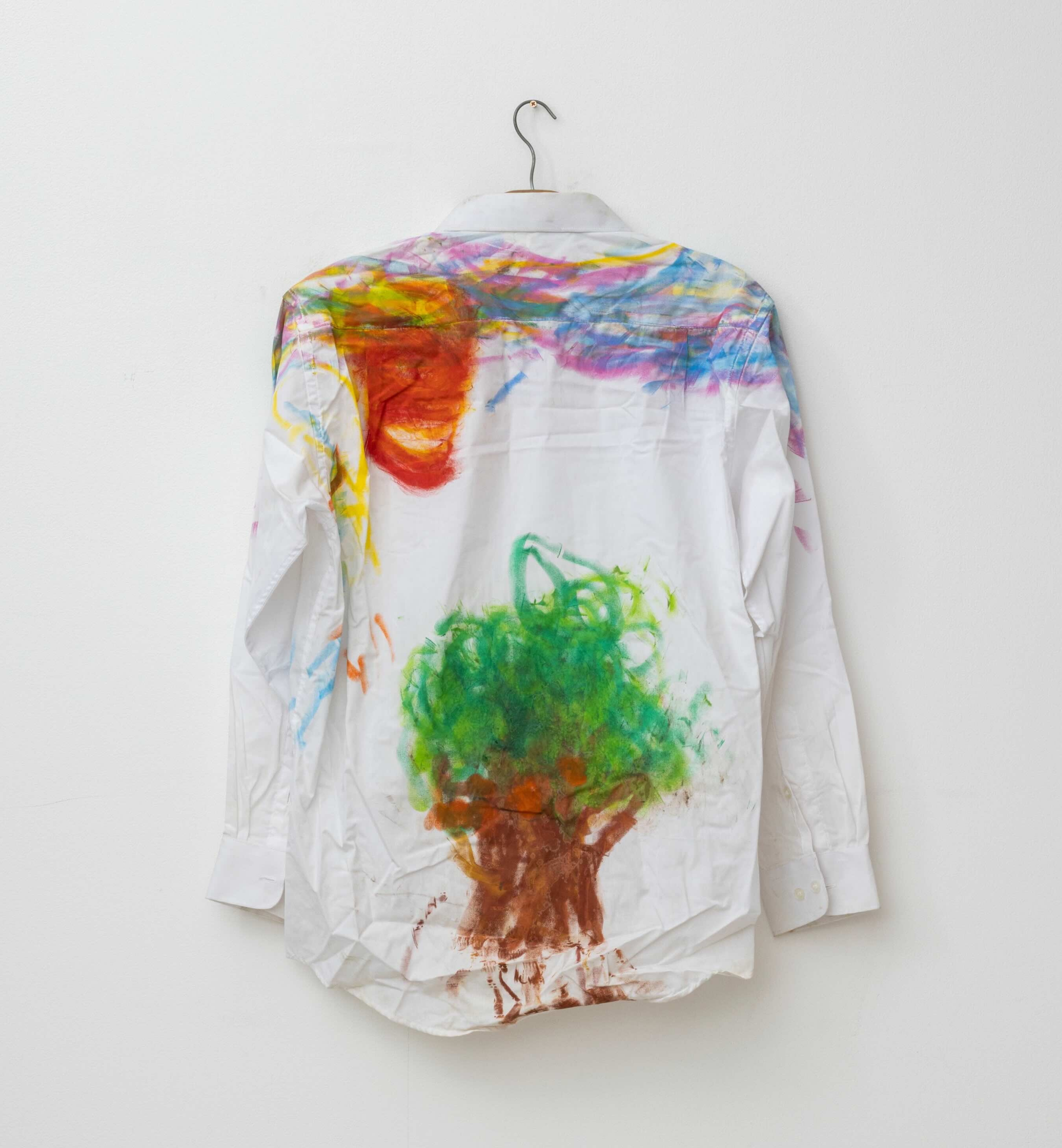 A white button-down shirt hangs on a wire hanger against a white wall. The back of the shirt is covered with colourful crayon, and the image at the bottom of the shirt recalls a tree.