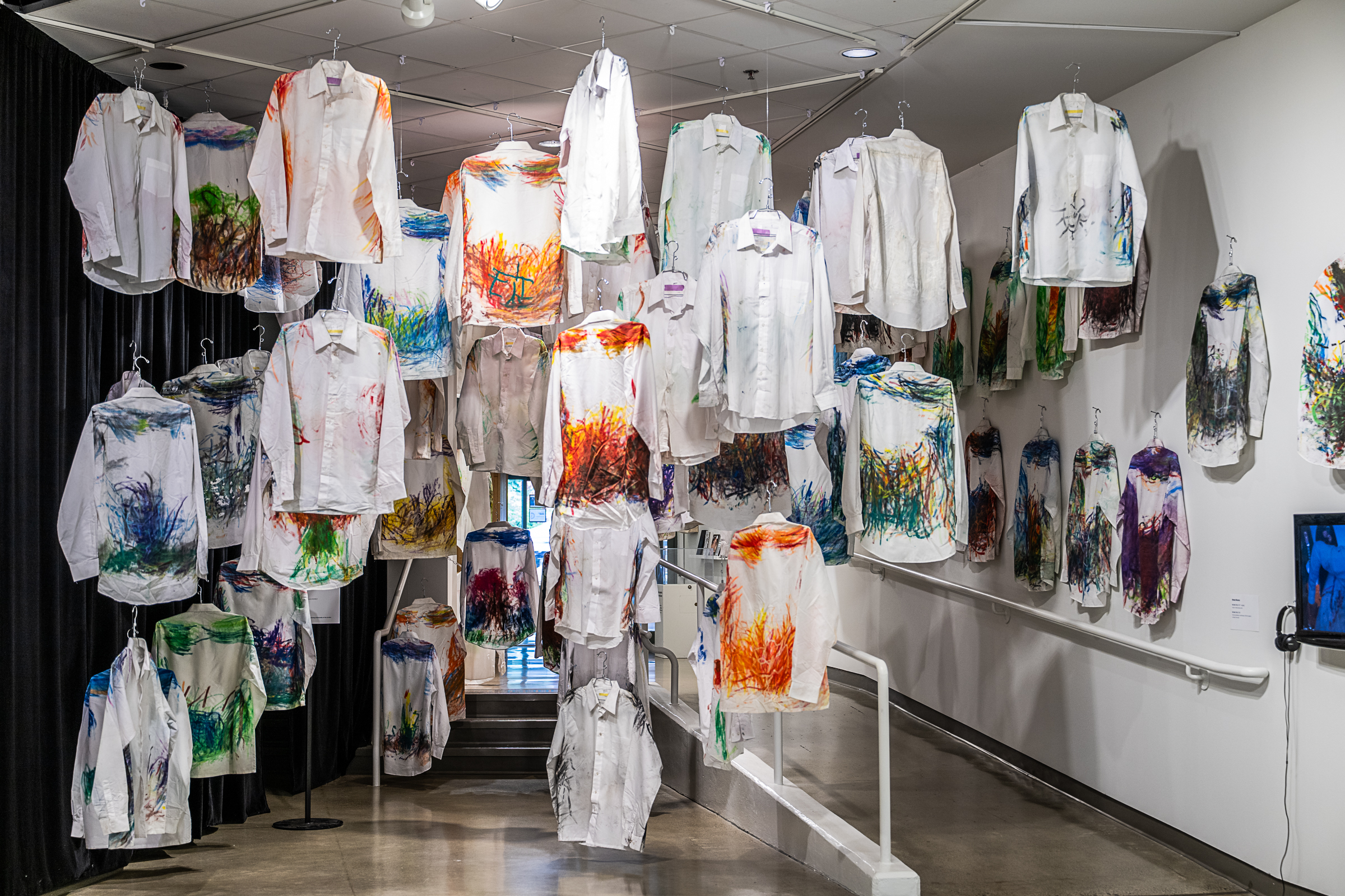 A large collection of white button-down shirts hangs on wire hangers in a gallery space, forming an informal archway and entrance into the gallery.