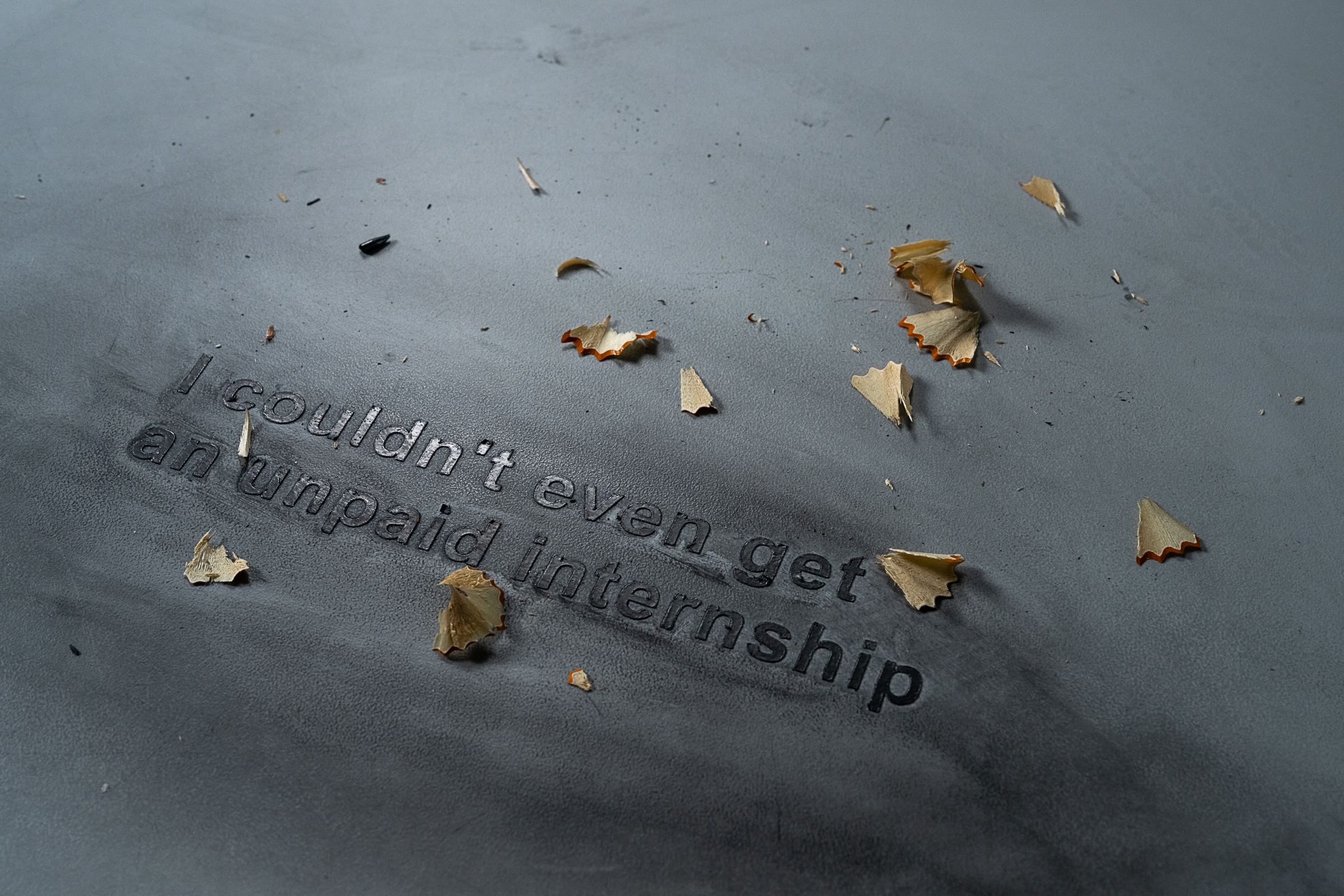 Pencil shavings are scattered across a matte flat dark grey surface. Text reads &ldquo;I couldn&rsquo;t even get an unpaid internship.&rdquo;