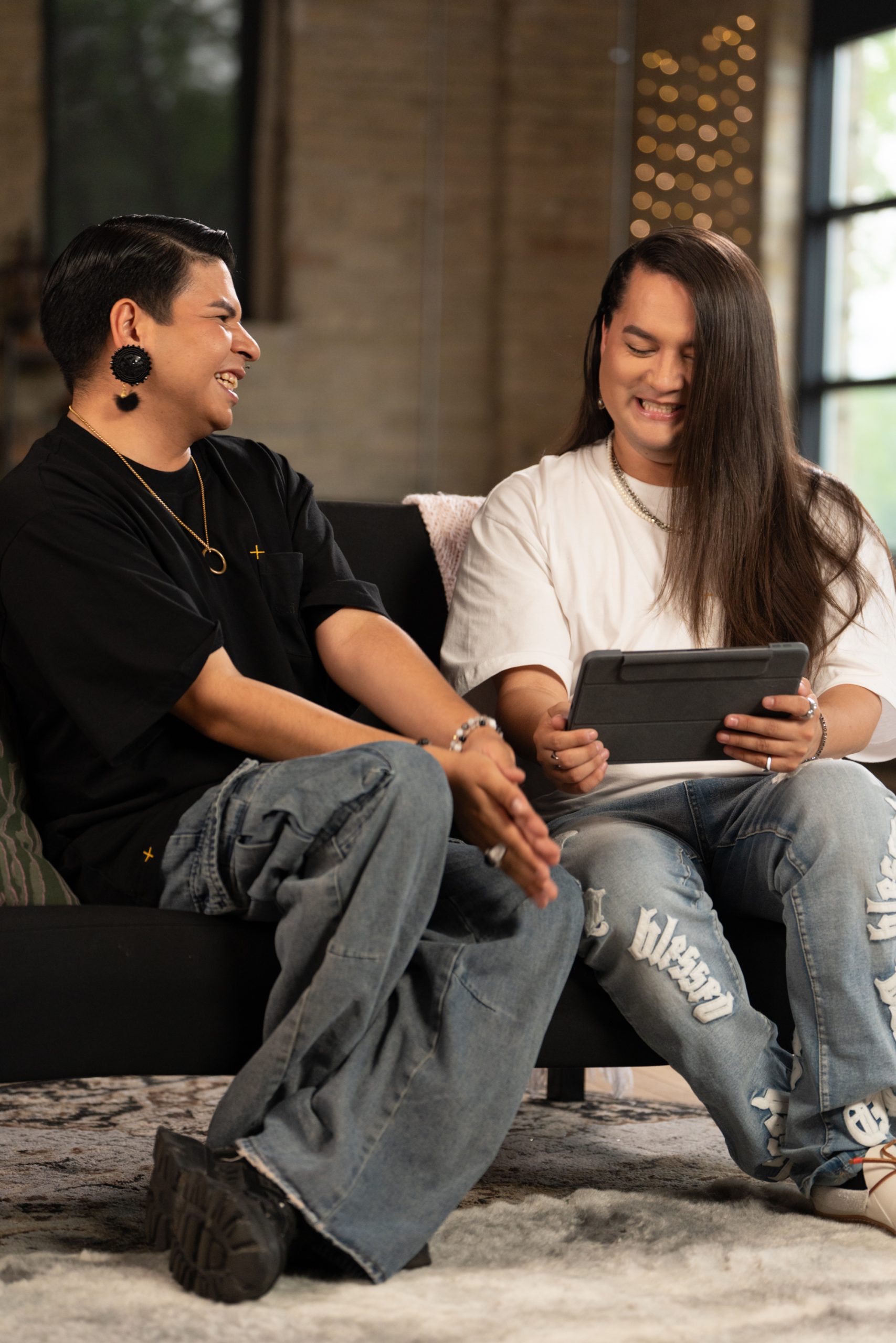 Scott Wabano, left, looks to Kairyn Potts, rights, and laughs. He is wearing a black T-shirt and jeans. Potts is wearing a white T-shirt and jeans and holding a tablet device.