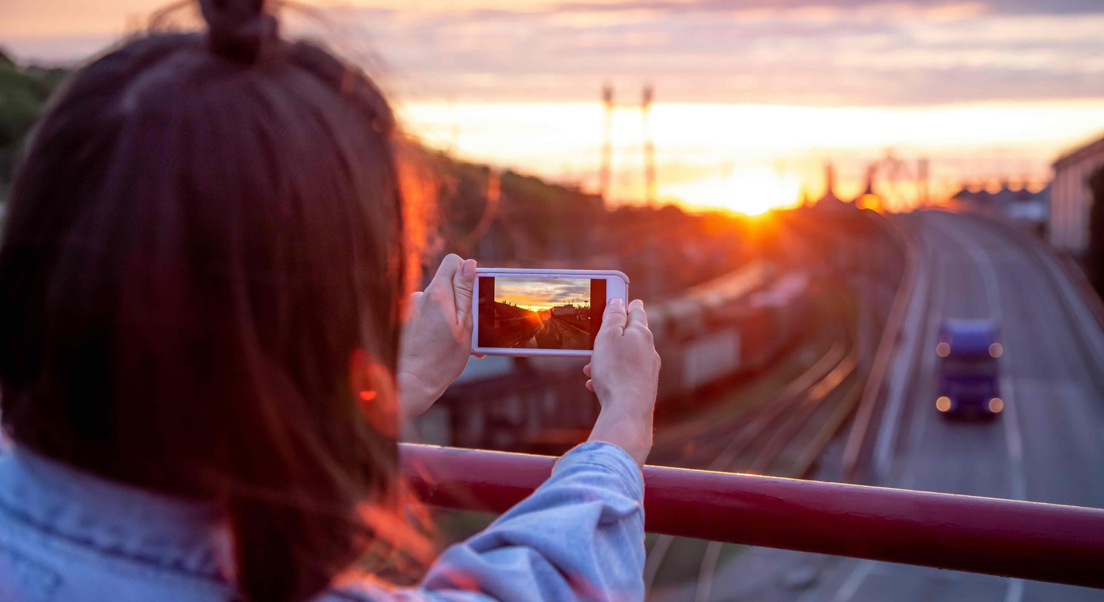 A young woman with long hair tied in a loose bun stands on a bridge, photographing the sunset and roadway below with her smartphone. She has her back turned to the camera and she is wearing a light denim shirt.