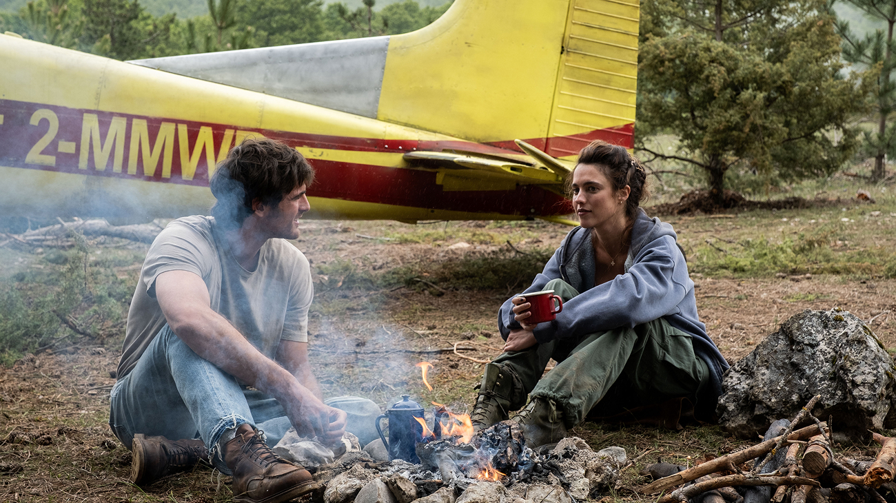 A man and a woman with brown hair in casual streetwear sit on the ground around a campfire encircled with rocks, mid-conversation. Behind them is a yellow plane in the forest.