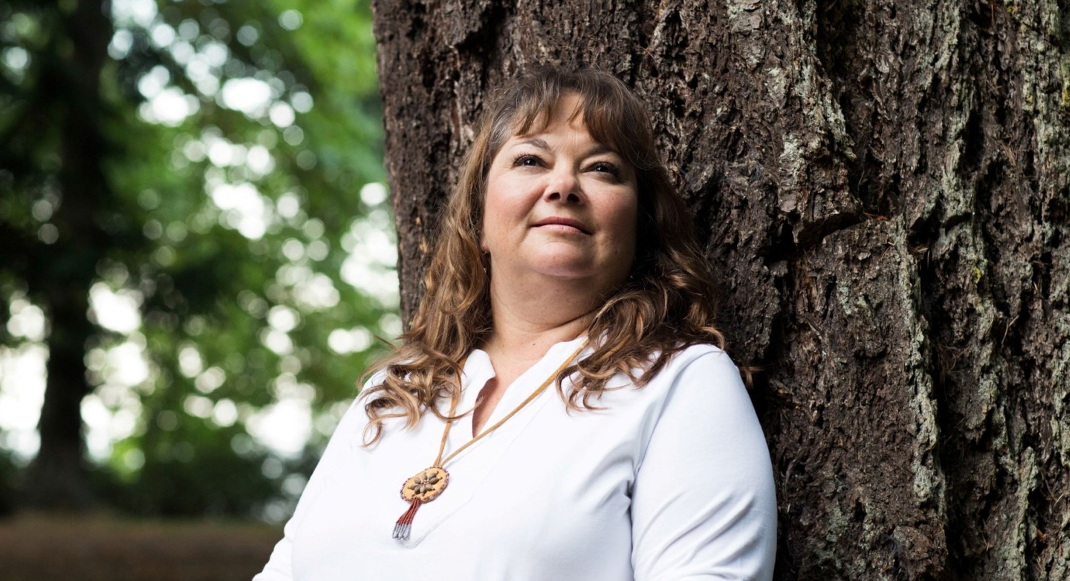 Monique Gray Smith has long curly brown hair. She is wearing a white top with a necklace featuring First Nations beadwork. She is standing against the thick trunk of a tree outdoors, looking to the middle distance.