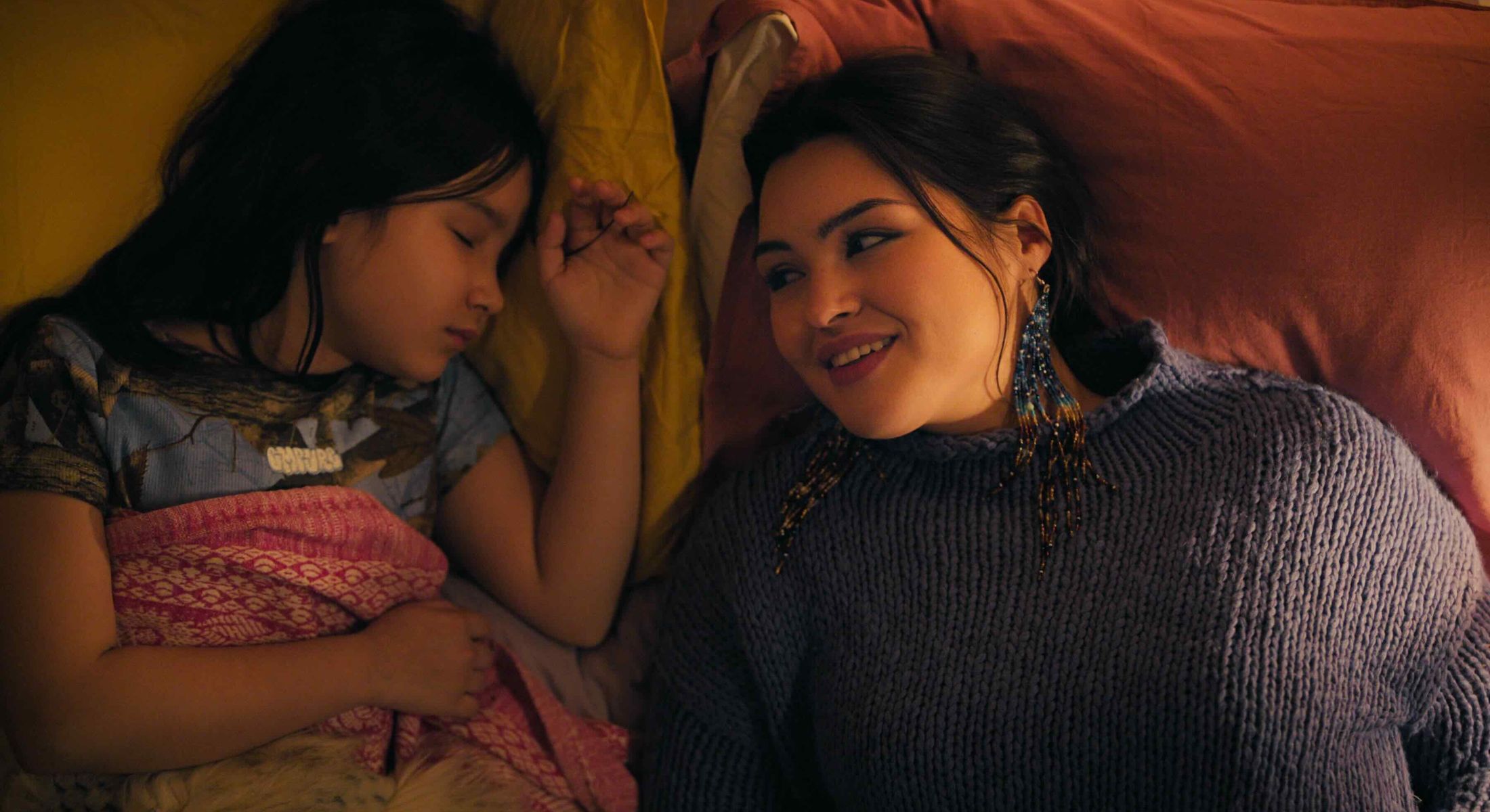 Two actors lie in bed together. On the left, Keira Cooper is a school-agd girl with long dark hair. She is asleep under a pink blanket. On the right, Anna Lambe has long dark hair pulled back. She wears colourful beaded earrings and a purple sweater. She is smiling at Cooper.