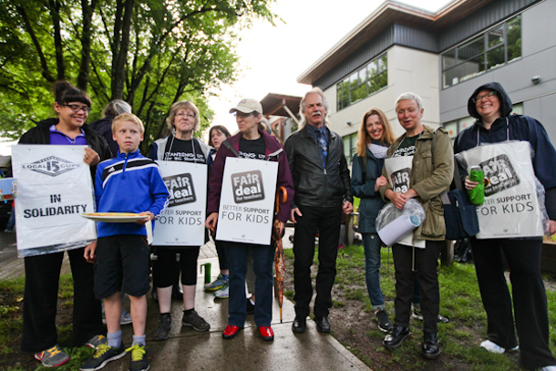 Ten people stand on a picket line, wearing rain gear and signs that read, “FAIR DEAL: Better Support for Kids.”