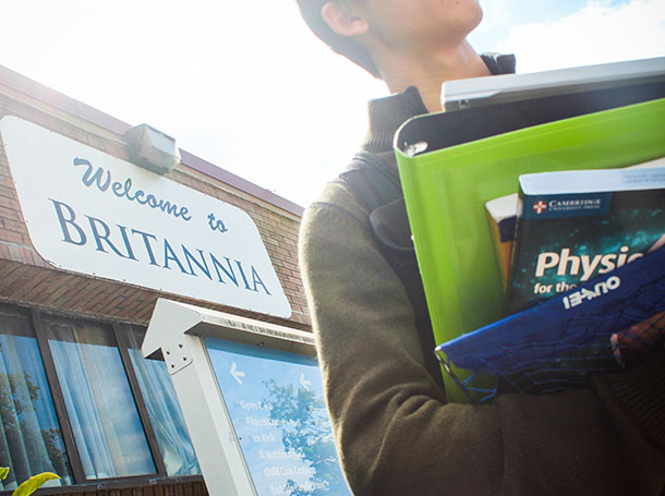 A student in an olive green sweater holds an armful of books in front of a white sign with navy text that reads &ldquo;Welcome to Britannia.&rdquo; We can&rsquo;t see the student&rsquo;s face. The sky is bright and white.