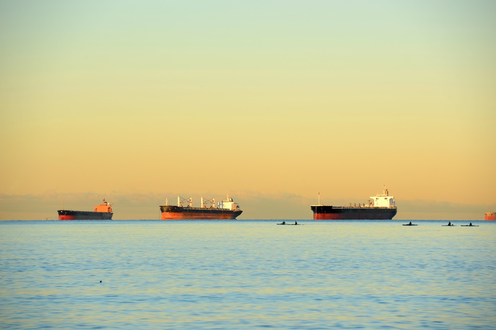 Three oil tankers are anchored off the coast of Vancouver. Kayakers are visible in and around them. 