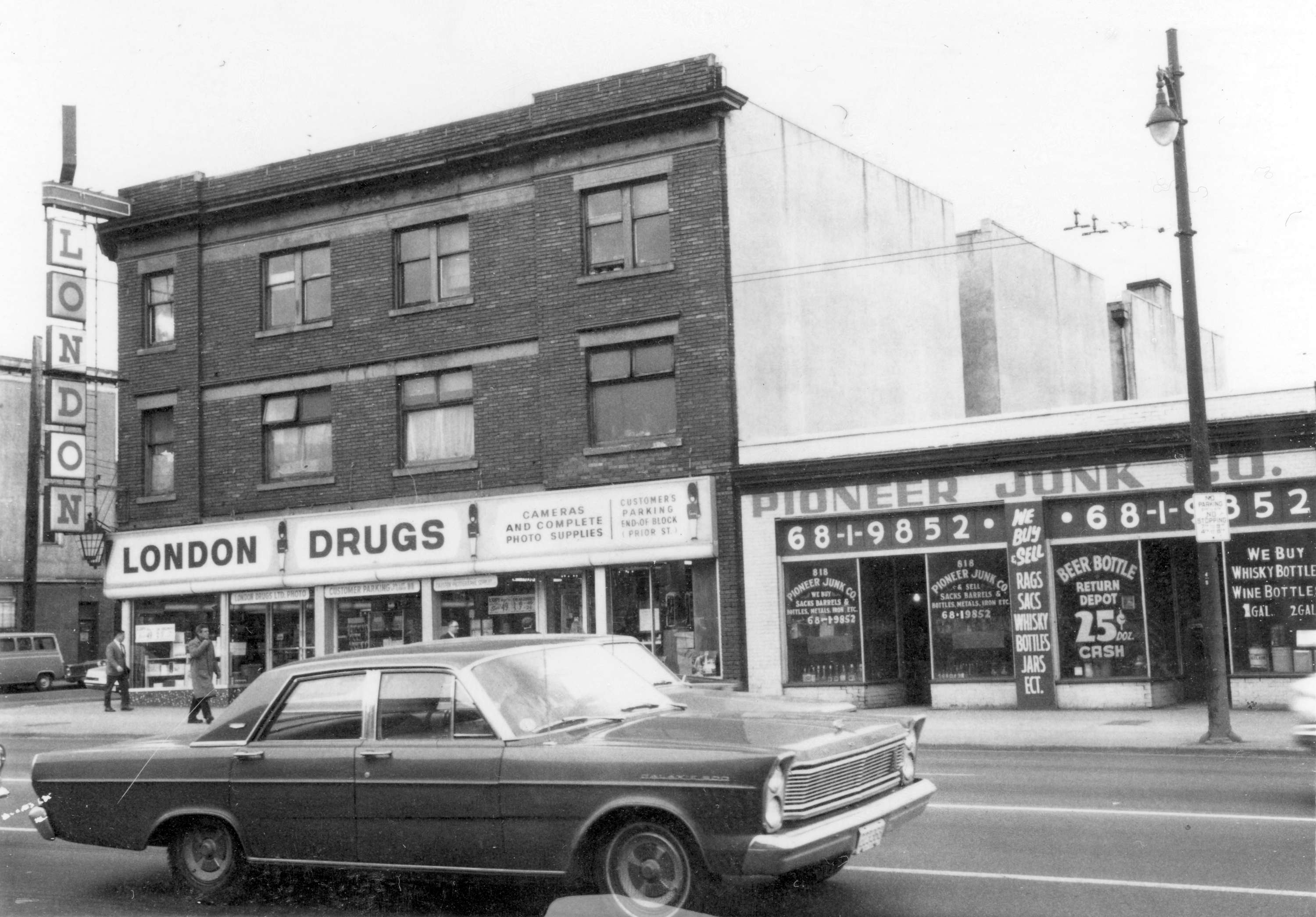 A black and white photo of Vancouver&rsquo;s main street, taken in 1968. On the right is a one-story building home to the Pioneer Junk Co., with painted signs on the windows advertising bottle returns for cash. On the left is a three-story building, with a London Drugs store on the main floor. A sign reads, &ldquo;Camera and complete photo supplies.&rdquo;