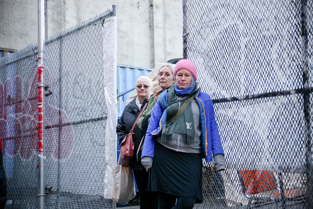 Three women stand together against chain-link fences in front of a grey concrete wall and light blue shipping container that house the fledgling Overdose Prevention Society in Vancouver&rsquo;s Downtown Eastside in 2016. Sarah Blyth is wearing a pink toque and blue jacket. Behind her are Overdose Prevention Society co-founders Ann Livingston (in a green jacket) and Chris Ewart (in dark glasses).