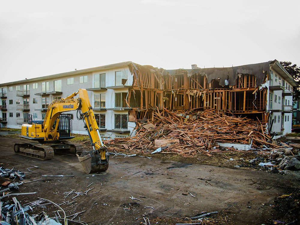 A digger beside an old walk-up apartment building with a chunk taken out of it, wood spilling onto the dirt.
