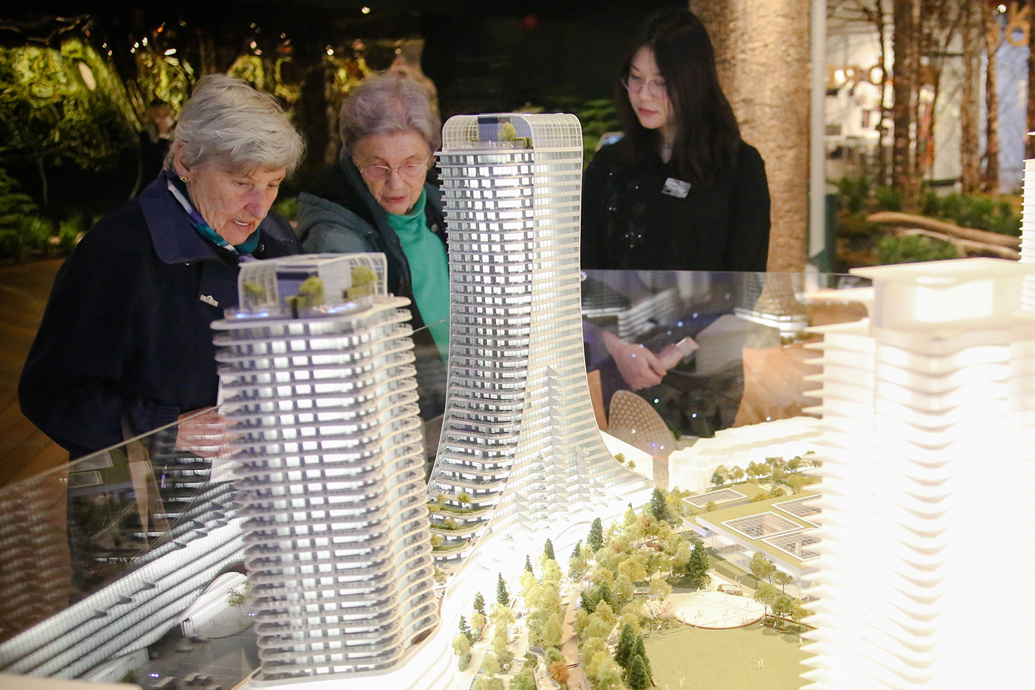 Two elderly women chat in front of a glowing architectural model of a large development&rsquo;s curvy towers.
