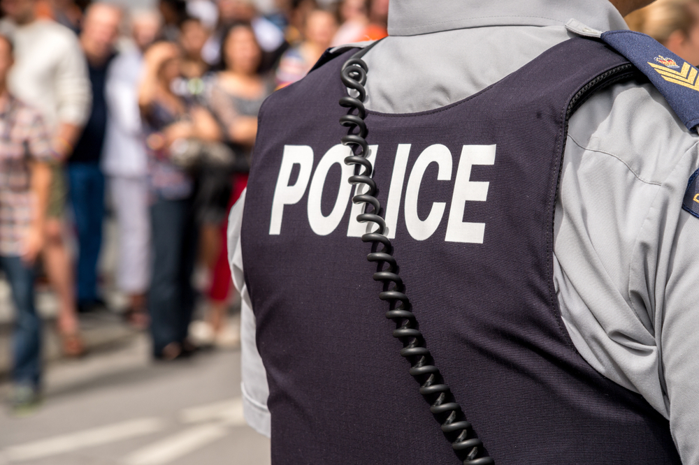 The back of a stab vest with the word ‘POLICE’ is worn by an officer standing amidst a crowd. Only their torso is visible.
