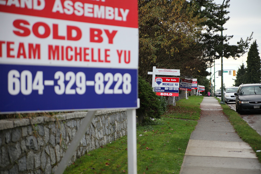 A chain of 'For Sale' signs stretches along a grey sidewalk in front of a half-dozen houses.