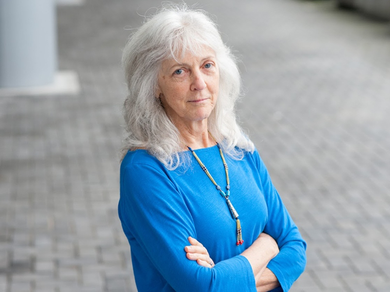 An older woman with shoulder-length grey hair and a light skin tone looks at the camera, wearing a blue long-sleeved shirt, her arms crossed.