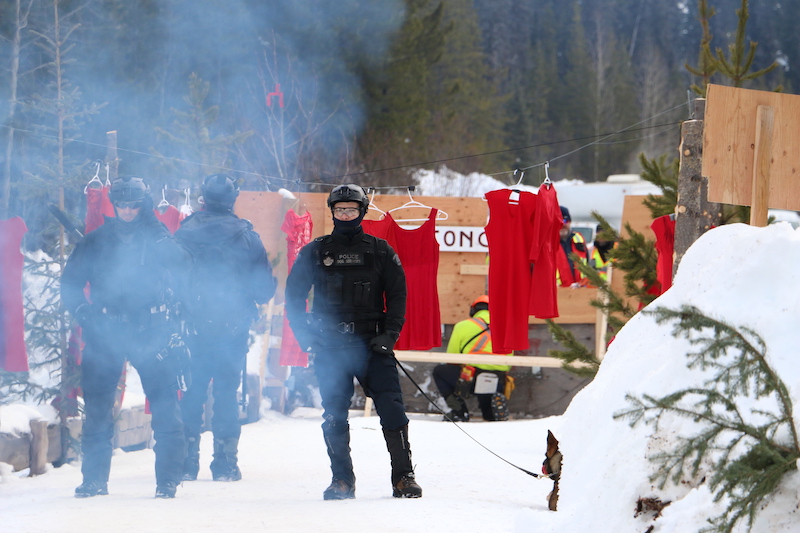 It is snowy. Three RCMP officers stand in uniforms with helmets and bulletproof vests. One officer has a dog. In the background, land defenders have placed red dresses on clothes hangers and strung them across the road.