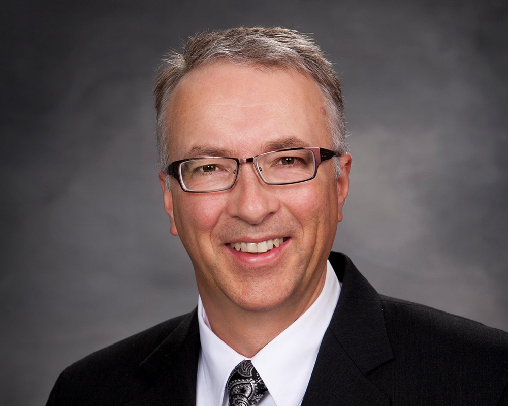 A light-skinned man with short grey hair and glasses and wearing a dark suit, white shirt and tie smiles at the camera.
