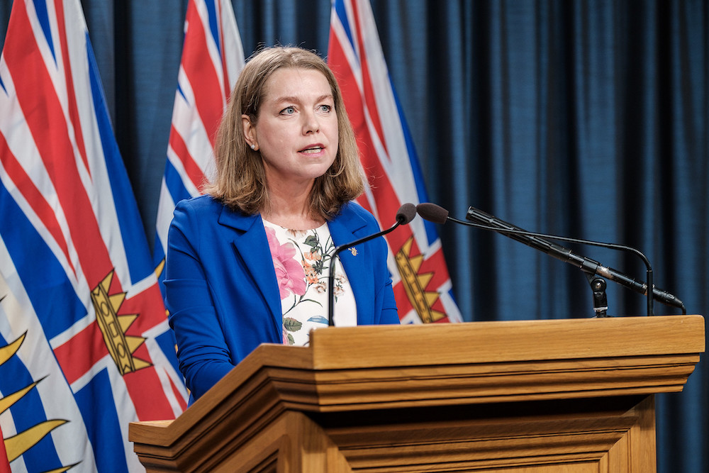 A woman with shoulder-length hair speaks at a podium, wearing a blue blazer.