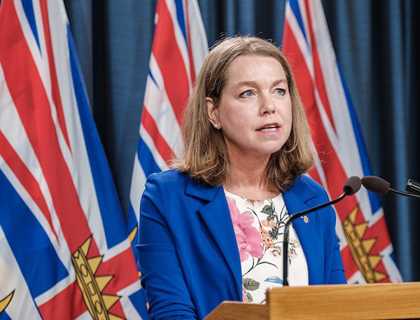 Mitzi Dean, a white woman with shoulder-length light brown hair, stands at a podium. She wears a medium blue jacket and flowered shirt. B.C. flags are behind her.