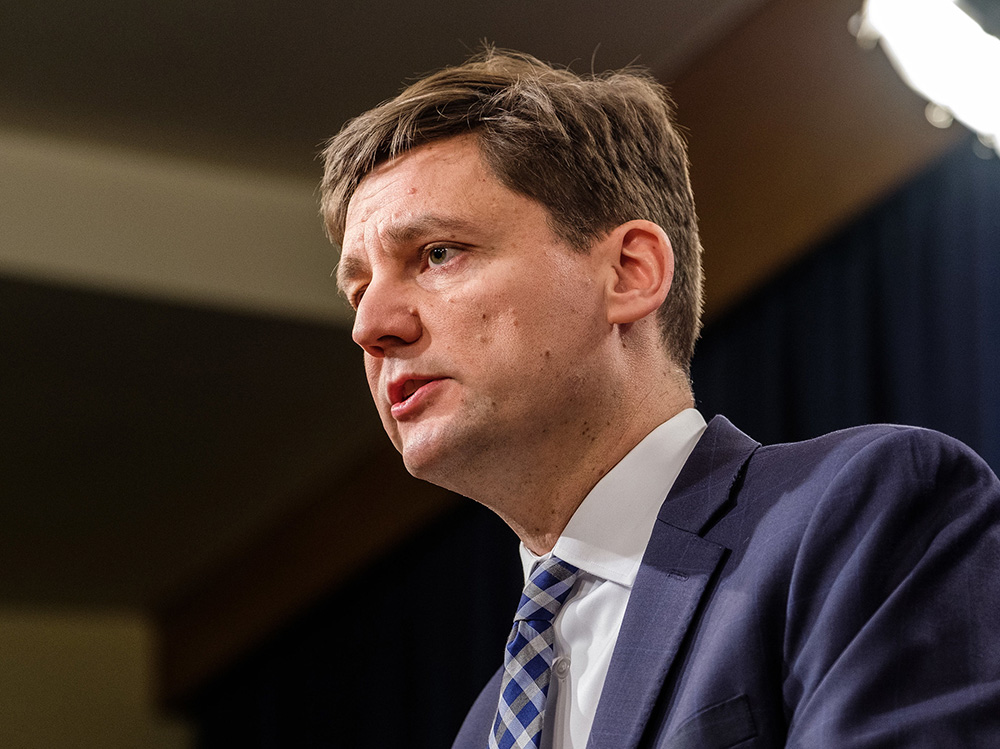 A portrait of David Eby in three-quarters profile. He&rsquo;s wearing a blue suit with a blue tie, and looks to be listening to someone off-camera.