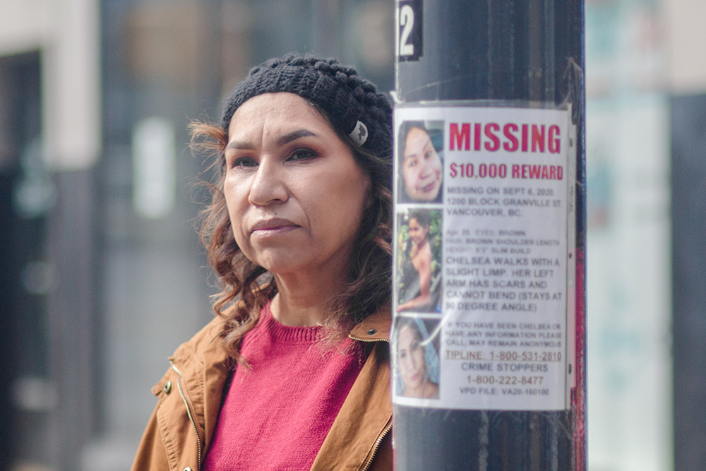 A woman, Sheila Poorman, stands next to a pole with a "Missing: $10,000 Reward" notice of her daughter, Chelsea Poorman.