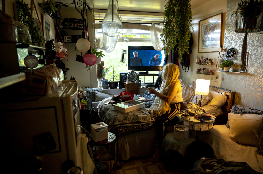 A woman with long blond hair sits on her bed in a small, well-taken-care-of room full of knick-knacks in an SRO. The window is open. She is watching 'Star Trek' on TV.