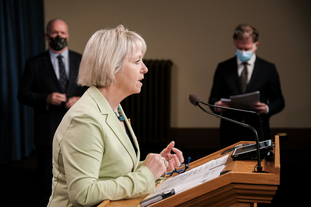 A light-skinned woman with cropped blond hair, wearing a pale green jacket, leans on a podium and speaks.