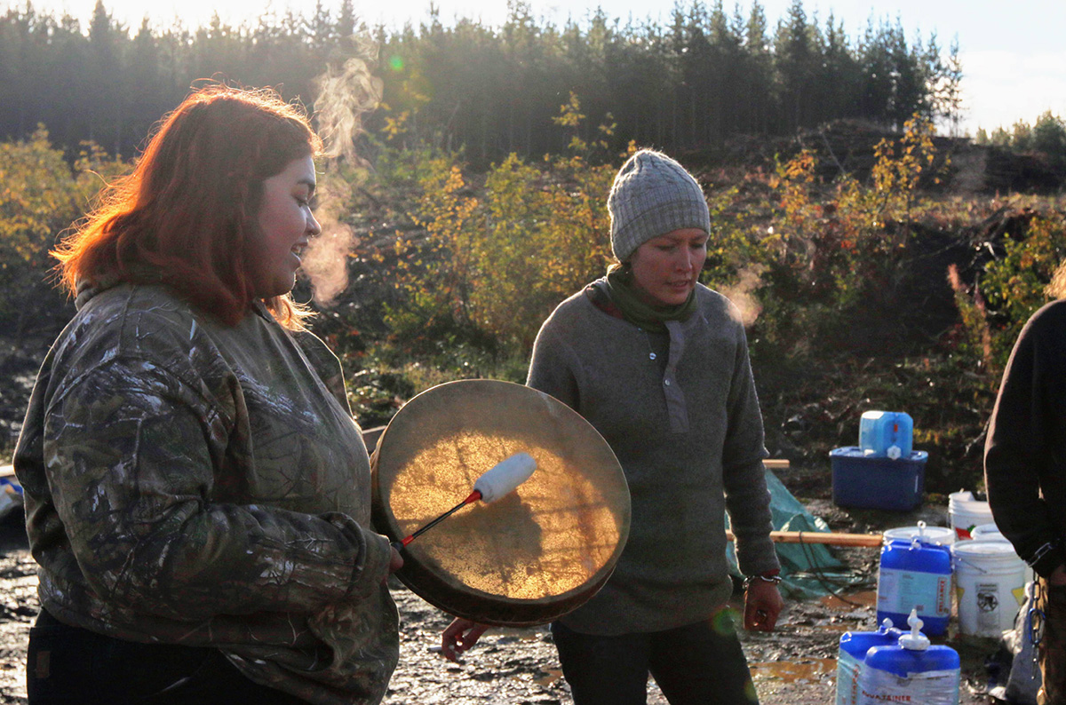 Two women stand together. One drums. It’s cold but not snowy; their breath is visible in the air.
