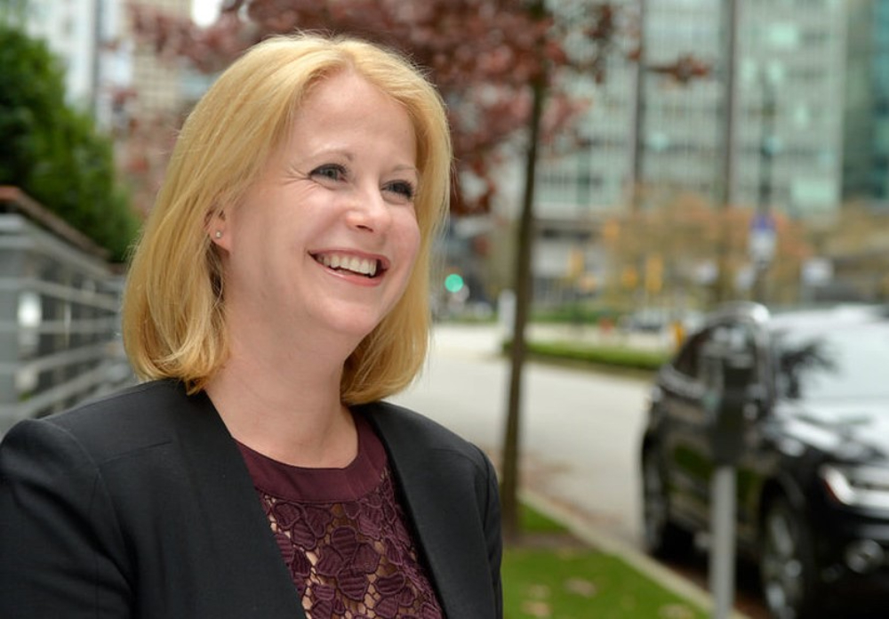 A woman with blond hair, wearing a dark jacket and purple short, smiles in closeup photo taken outdoors.
