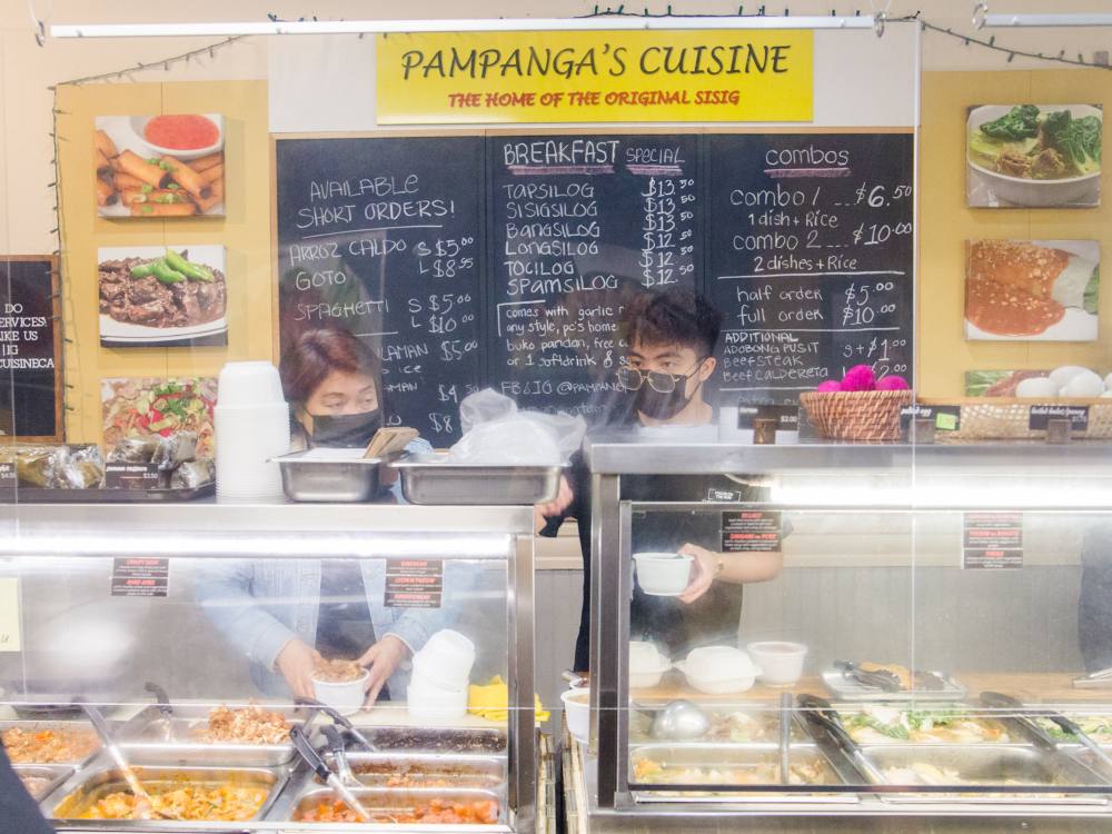 Edith Malang, left, and her son J, right, serve food from behind the counter at Pampanga&rsquo;s Cuisine, a Filipino restaurant on Joyce Street in Vancouver. Both are wearing black medical masks. Edith is placing a full takeout container on the counter in front of her while J consults an order receipt as he packages a variety of meal items. In front of them, food is in stainless-steel warming trays. Behind them, menu items are handwritten on a chalkboard. 