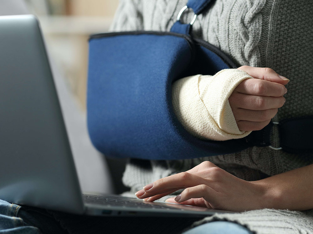 A person with a broken arm in a sling uses a laptop touch pad with their free hand.