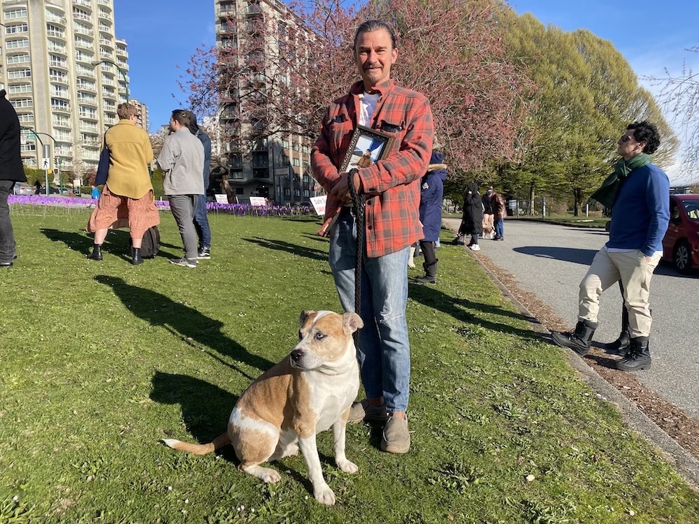 Matthew Witt, clad in a checkered red and black flannel shirt and jeans, stands in a grassy field holding a picture of his late son Sebastian. His dog Rio sits before him. Other people stand around him facing the rows of purple flags in the background.