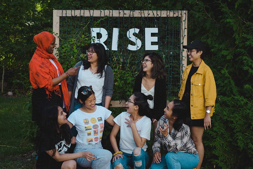 A group of eight young racialized people are gathered in front of a handmade sign that reads &ldquo;Rise.&rdquo; They are the founders of Vancouver&rsquo;s Shades of Sustainability, which aims to create space for dialogue about environmental practices among BIPOC people. Members of the group are looking at each other, smiling and laughing.