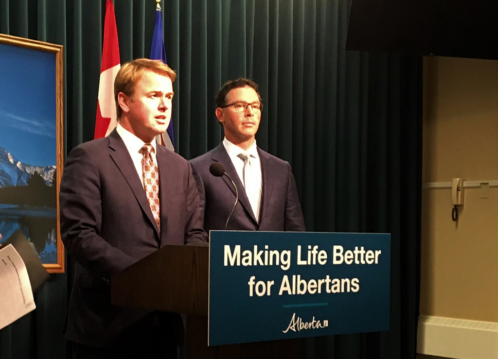 Two people, government officials, clad in suits stand before a podium appearing to address questions.