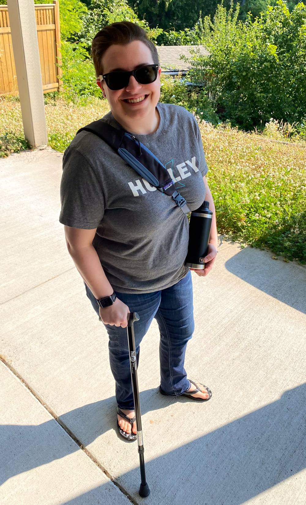 Britt Helm stands in the sun with a black cane in their right hand. They are wearing sunglasses and smiling.