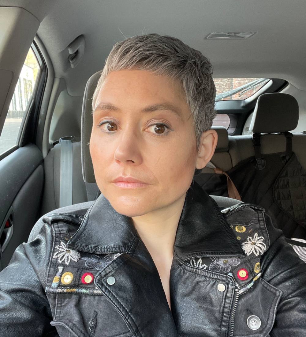 Jaigris Hodson looks at the camera while sitting in their car. Their hair is short and blonde, and they are wearing a leather jacket with embroidery on the front.