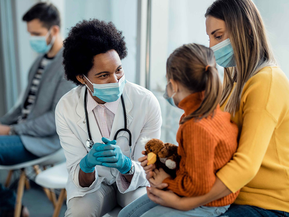 A doctor wearing a white jacket, with stethoscope and face mask, speaks with a young girl who is seated on her mother&rsquo;s lap holding a stuffed animal toy.