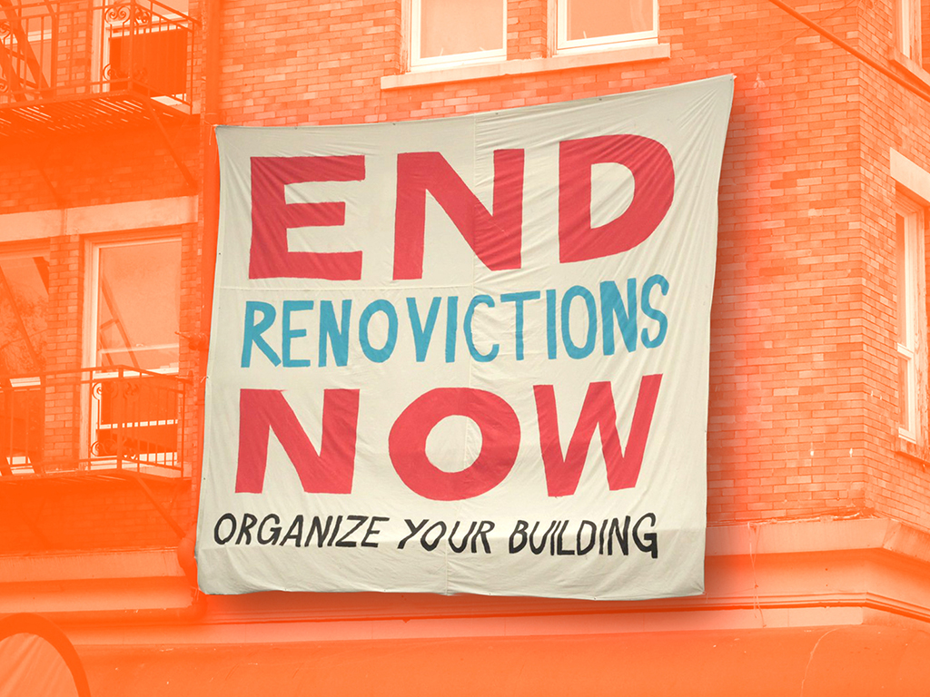 A banner reading &ldquo;End Renovictions Now. Organize Your Building&rdquo; is stretched across a brick apartment building.