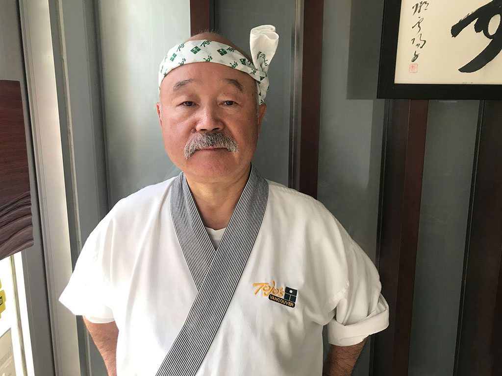 Chef Tojo wears a Tojo&rsquo;s Vancouver chef&rsquo;s coat and headband. He has a salt-and-pepper mustache.