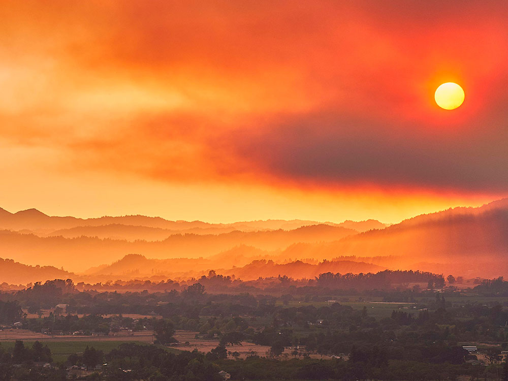 The Californian landscape, covered with trees and hills, as wildfire smoke blots out the sun and paints the horizon a deep orange hue.