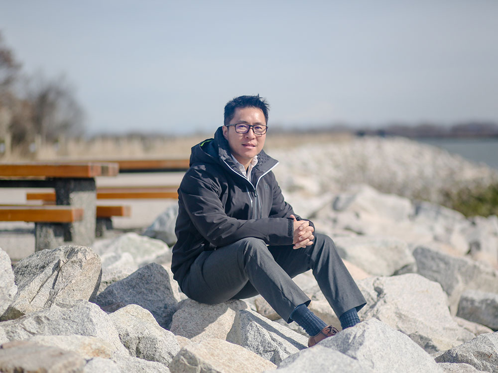 Jason Ho sits on a pile of large boulders that slope down to the Fraser River on a sunny day.
