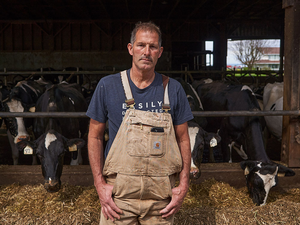 A man in tan overalls and a navy blue t-shirt stands in front of a barn of black and white milking cows at his family&rsquo;s farm in Pitt Meadows, BC.