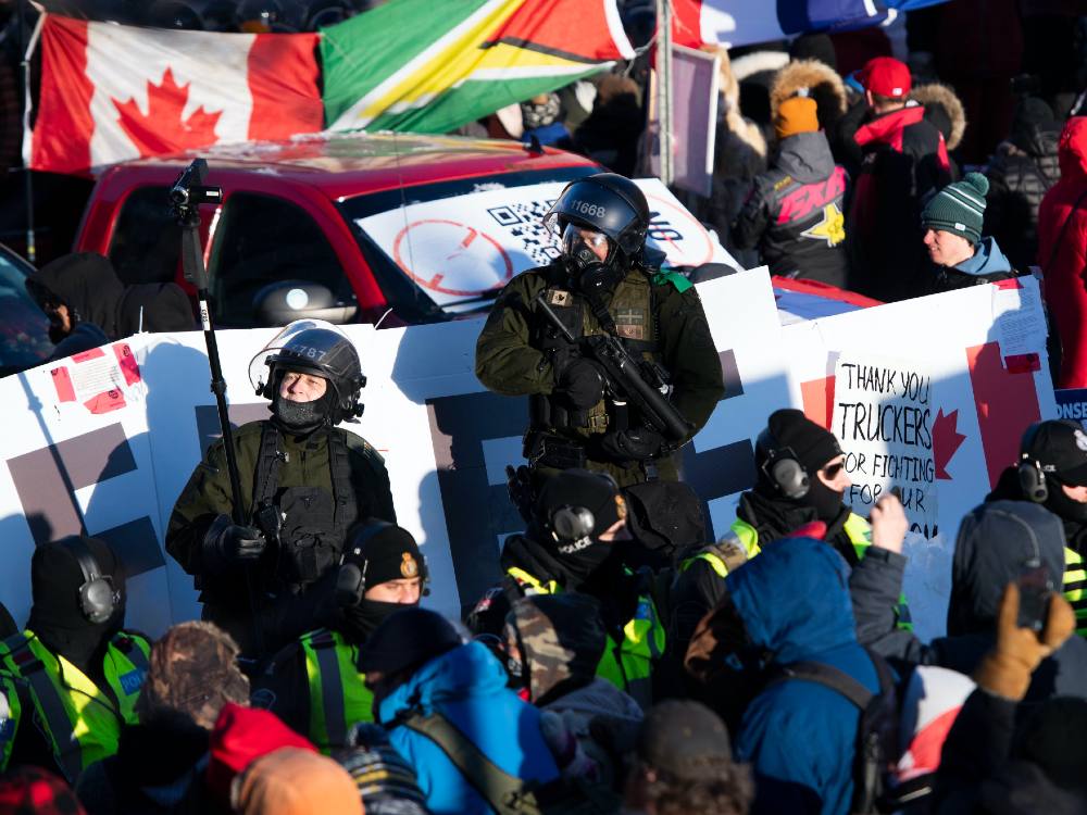 Two riot police stand in the middle of a packed crowd at the Freedom Convoy protests in February 2022. One holds a gun, the other holds a video camera attached to a pole. A sign beside them reads "Thank you truckers." 