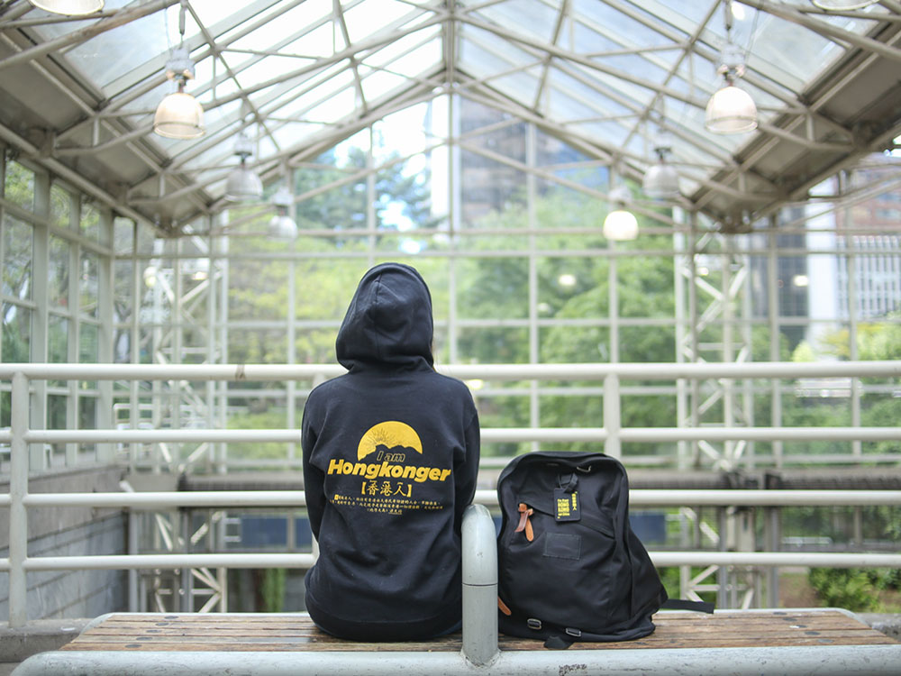A woman sits on a bench at a transit station with her back to the camera. She&rsquo;s wearing a black hoodie, with yellow text on the back that says &lsquo;I am Hongkonger&rsquo; in English and &lsquo;Hongkonger&rsquo; in Chinese characters. She has a black backpack beside her with a tag that says, &lsquo;I&rsquo;m from Hong Kong, not China.&rsquo;