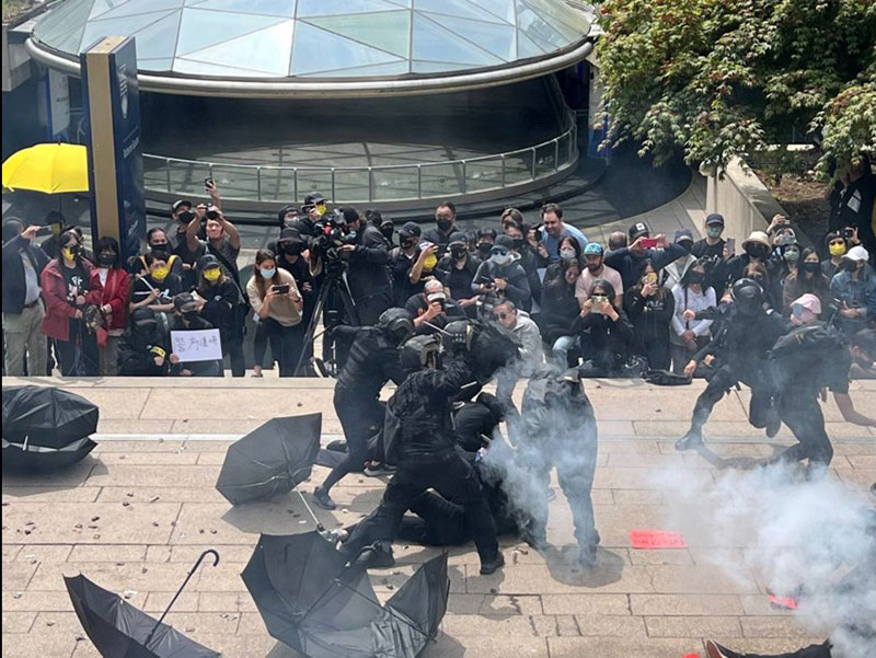 A crowd watches the re-enactment of 2019&rsquo;s protests in Hong Kong in a downtown plaza. There are umbrellas scattered on the ground as actors recreate a chaotic scene of police beating protesters with batons.
