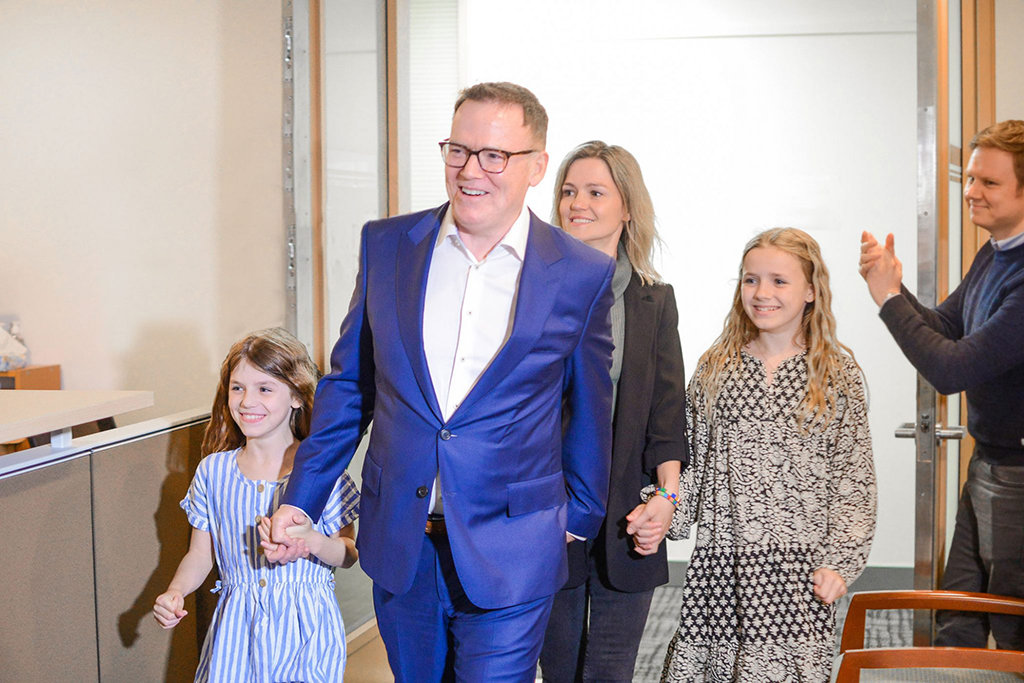 Kevin Falcon is wearing a blue suit and glasses. He is smiling and walking through a doorway into a room of supporters after winning the Vancouver Quilchena by-election in April 2022. He is holding hands with his youngest daughter, who is wearing a blue and white striped dress. Falcon&rsquo;s spouse and oldest daughter walk behind him.