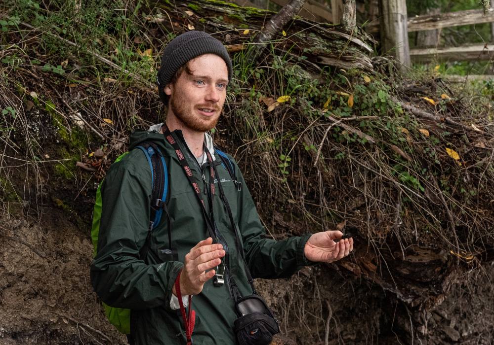 A person wearing a green rain jacket and a whistle and a tuque stands with their palms up in front of a greenery.