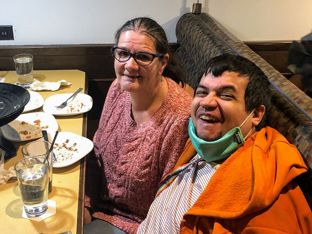 A woman and a man sit at a restaurant table, both smiling. He wears an orange jacket and COVID mask around his neck. She has glasses and a pink shirt.